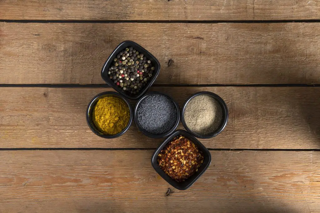 Five small bowls with mix of peppers, white pepper, red dry chillies, puppy seeds and curry on a wooden surface