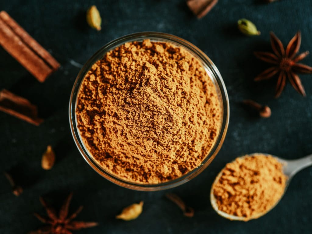 Indian or pakistani masala powder in spoon and small glass bowl. Close up view of homemade dry curry garam masala mix spices blend on dark background.
