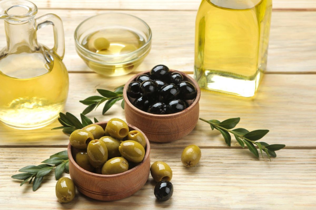 Green and black olives in a wooden bowl with leaves and olive oil on a natural wooden table.