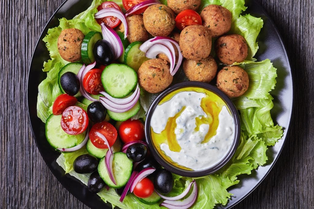 Mediterranean platter of falafels, tomatoes, cucumber, red onion, black olives, on the fresh salad leaves served with tzatziki sauce served on a black plate on a dark wooden background, close-up