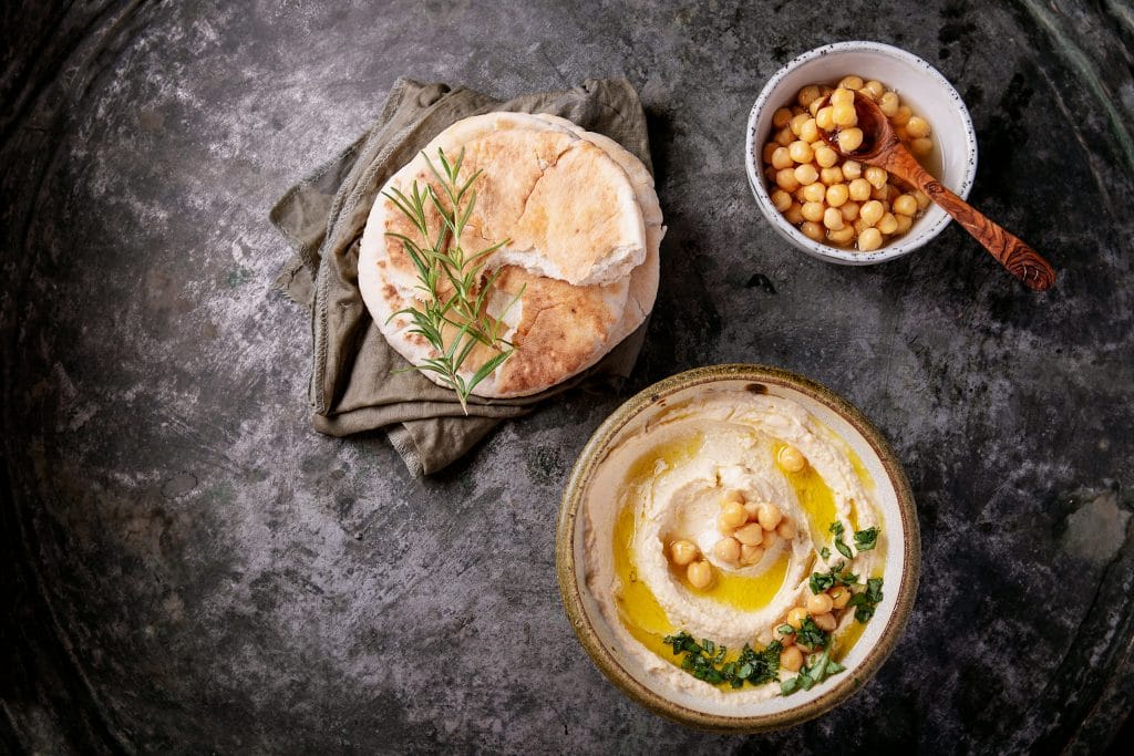 Home made hummus bowl, decorated with boiled chickpeas, herbs, pita and olive oil over a rustic metal background. Top View.