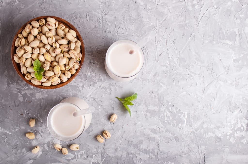Organic non dairy pistachio milk in glass and wooden plate with pistachionuts on a gray concrete background. Vegan healthy food concept, flat lay, top view, copy space.