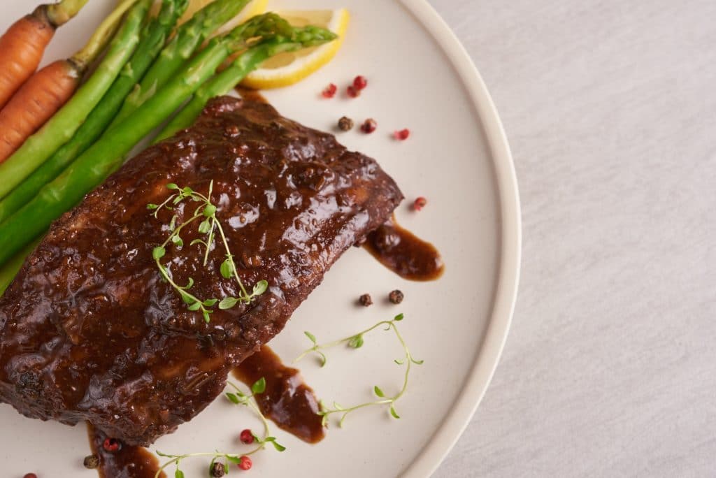 Pork roasted, grilled spare ribs from a summer BBQ served with Vegetables, asparagus, baby carrots, fresh tomatoes and  spices. smoked ribs in white plate on stone background . top view.