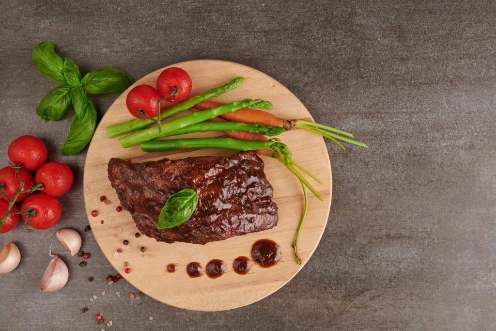 Pork roasted, grilled spare ribs from a summer BBQ served with Vegetables, asparagus, baby carrots, fresh tomatoes and  spices. smoked ribs on wooden cutting board  on stone background . top view.