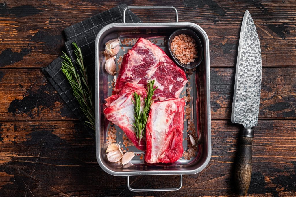 Raw beef Short Ribs in steel tray with rosemary. Wooden background. Top view.