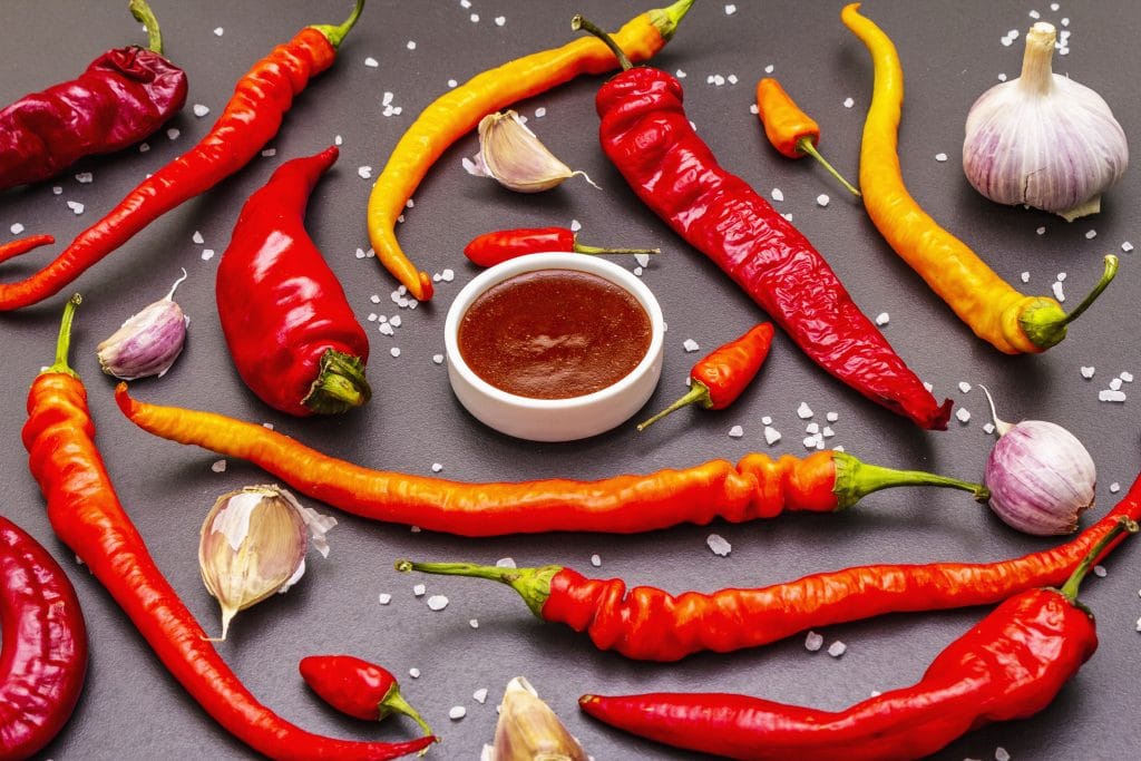 Traditional sauce Sriracha with ingredients. Red, orange and yellow chilli, garlic, sea salt. In ceramic bowl, black stone concrete background, close up