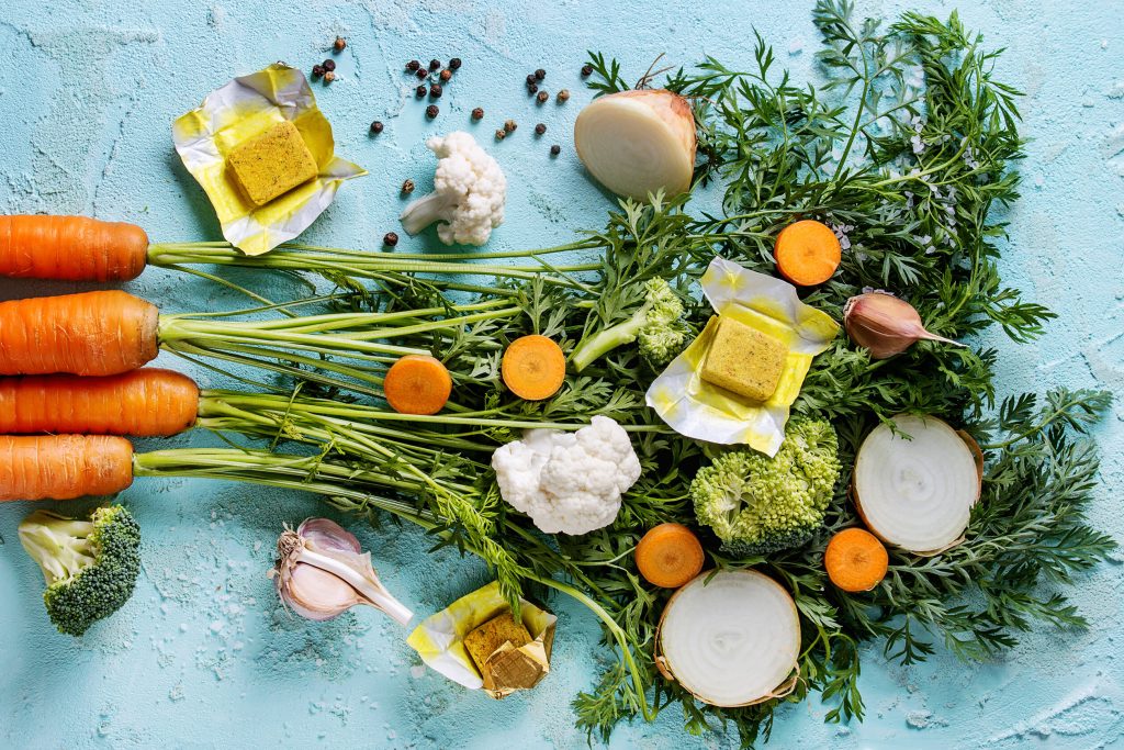 Raw vegetables and bouillon cubes for cooking soup. Young carrot with haulm, broccoli, cauliflower, onion, garlic, salt pepper over turquoise concrete background. Top view. Dinner cooking concept