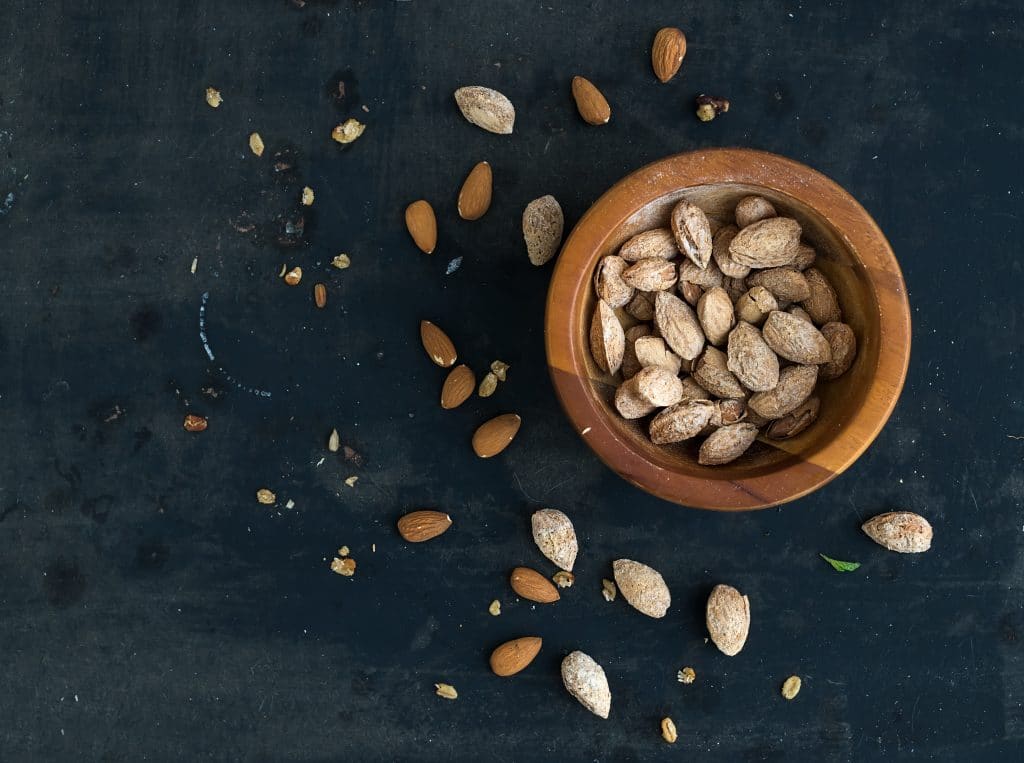 Wooden bowl of salty almond nuts in nutshell grunge dark backdrop, top view, copy space