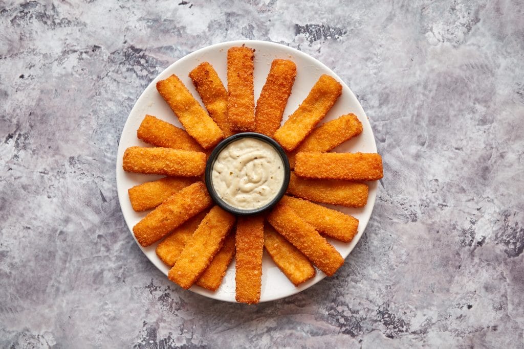 Crumbed fish sticks served with garlic dip sauce on a white plate on a stone table. Top view with copy space.